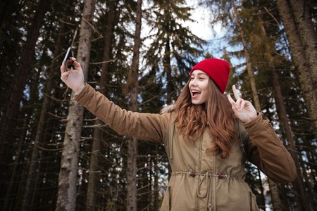Photo of young happy woman standing over forest and looking at phone while make a selfie and gesturing with hands.の写真素材