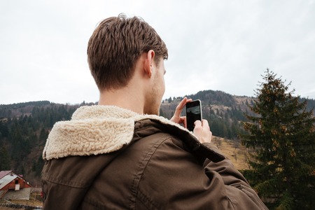 Back view of handsome young man take a photo by his phone in the mountains.の写真素材