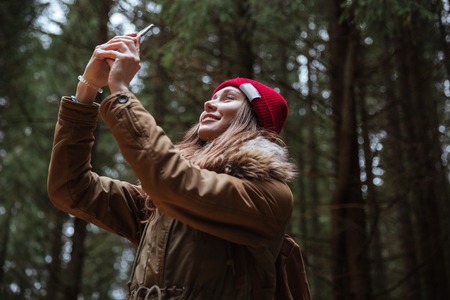 Image of cheerful young lady standing in the forest make a photo by phone. Looking aside.の写真素材