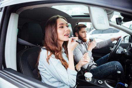 Pretty couple in car with coffee. man at the wheel. Girl paints her lipsの写真素材