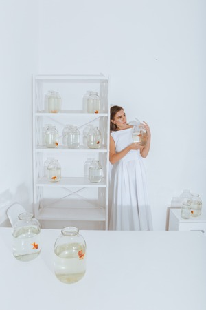 Cute young woman standing in the room and holding gold fish in jar