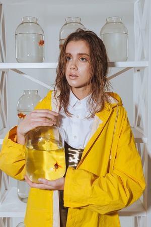 Pensive cute young woman in yellow raincoat holding jar with gold fish