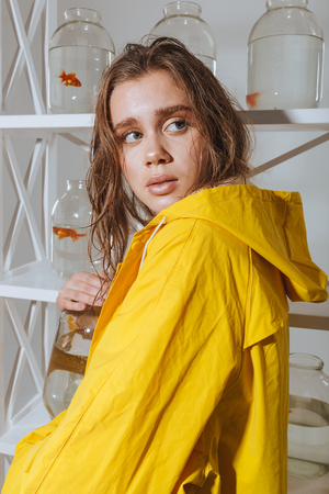 Scared young woman in yellow raincoat standing and hiding jar with gold fishの写真素材