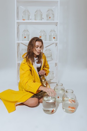 Lovely young woman in yellow raincoat sitting on the floor near jars with gold fishesの写真素材