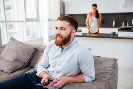 Man with girl on background. man sitting on bed and watching TV. woman cooked in kitchenの写真素材