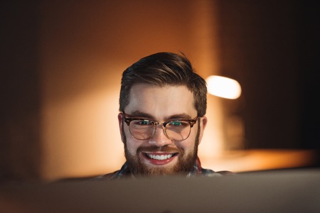 Image of bearded young happy web designer dressed in shirt in a cage print and wearing glasses working late at night and looking at computer.の写真素材
