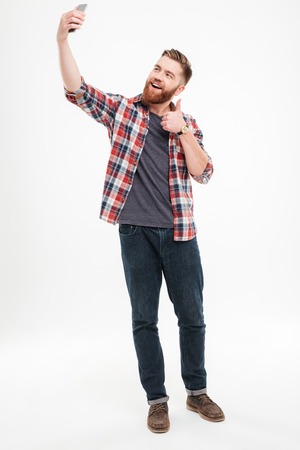 Full length portrait of a smiling casual man taking selfie and showing thumbs up gesture over white backgroundの写真素材
