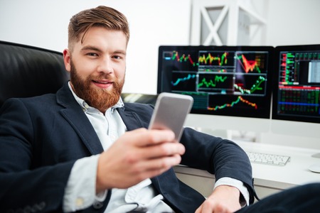 Happy bearded young businessman sitting and using mobile phone in office