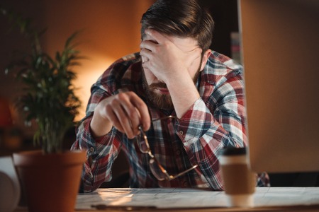 Photo of young confused bearded web designer dressed in shirt in a cage print working late at night...