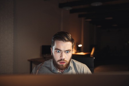 Shocked web designer dressed in shirt and holding eyeglasses working late at night and looking at computer.の写真素材