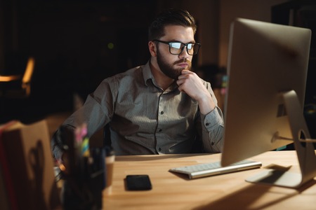 Photo of concentrated bearded web designer dressed in shirt working late at night and looking at...