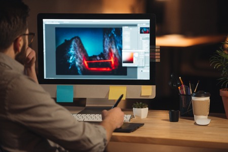Young concentrated bearded web designer dressed in shirt working late at night and looking at...
