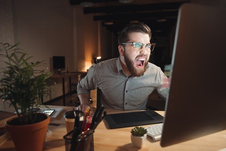 Picture of angry web designer dressed in shirt and wearing eyeglasses working late at night and looking at computer while screaming.の写真素材