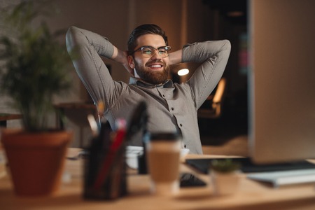 Picture of happy bearded designer dressed in shirt working late at night with computer and stretching.の写真素材