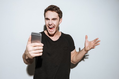 Image of young angry man dressed in black t-shirt standing over white background and using phone.の写真素材
