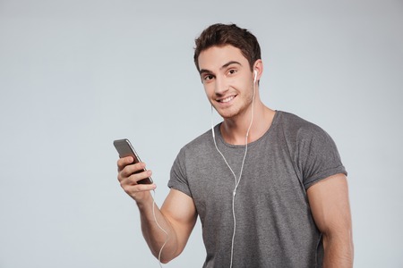 Portrait of a handsome casual man with earphones holding mobile phone isolated on the white...