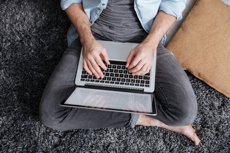 Top view of a casual man sitting on carpet and typing on laptop computer at homeの写真素材