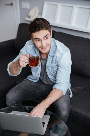 Portrait of a smiling satisfied man drinking tea while working on laptop on a sofa at homeの写真素材