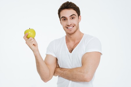 Photo of young cheerful man dressed in white t-shirt standing over white background showing alarm to camera holding apple.の写真素材