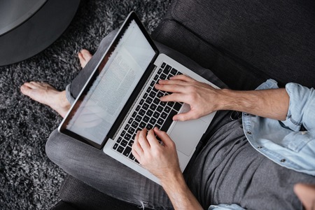 Top view of a casual young man typing on laptop while sitting on sofa home