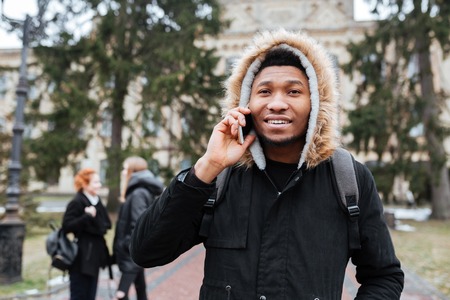 Portrait of a young african man student in jacket talking on mobile phone while standing at the university campusの写真素材