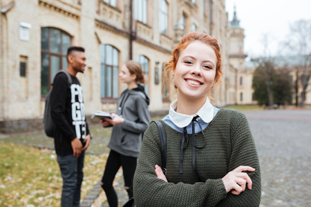 Portrait of a smiling redhead female student standing at the university campus with hands foldedの写真素材