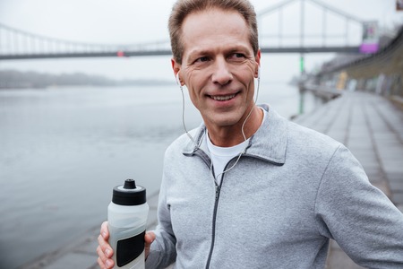 Close up runner in gray sportswear standing near the river with bottle of water in hand and looking awayの写真素材