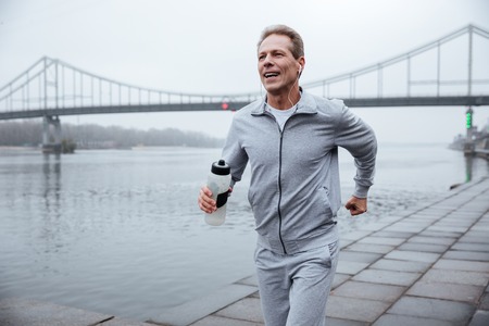 Man in gray sportswear running with bottle of water near the riverの写真素材