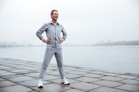 Full length Runner in gray sportswear standing with arms at hips near the water. Side view