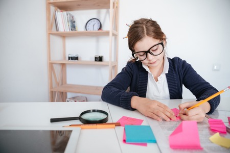 Concentrated little girl in glasses sitting and writing at the table