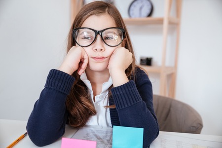 Sad bored little girl in glasses sitting at the table in classroomの写真素材