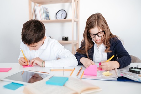 Concentrated little boy and girl sitting and writing on colorful sticky notes at the table
