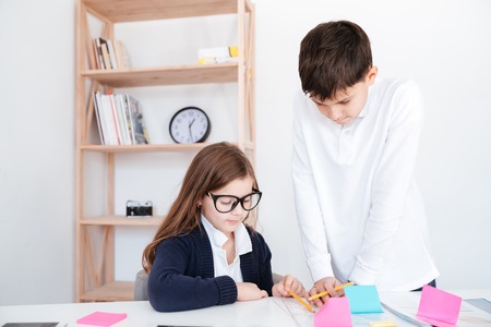 Boy standing and teaching little girl in glasses at the tableの写真素材