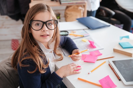 Happy little girl in glasses sitting and writing at the tableの写真素材