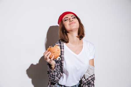 Picture of hungry young woman dressed in shirt in a cage print wearing hat standing isolated over white background and eating burgerの写真素材