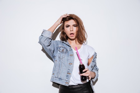 Picture of a young lady dressed in jeans jacket standing isolated over white background while drinking aerated sweet water.の写真素材
