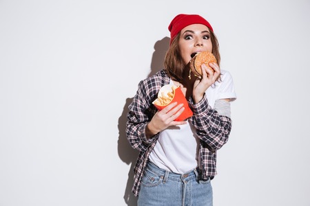Photo of hungry young woman dressed in shirt in a cage print wearing hat standing isolated over white background and holding fries and burgerの写真素材