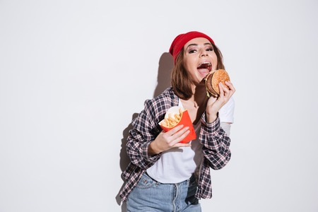Image of hungry emotional young woman dressed in shirt in a cage print wearing hat standing isolated over white background and holding fries and burgerの写真素材