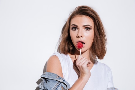 Picture of a young pretty lady dressed in jeans jacket standing isolated over white background while eating candy.の写真素材