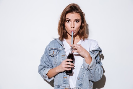 Photo of a young lady dressed in jeans jacket standing isolated over white background while drinking aerated sweet water.の写真素材