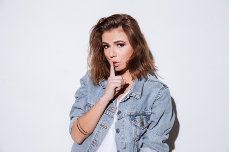 Image of a young lady dressed in jeans jacket standing isolated over white background make silence gesture. Look at camera.の写真素材