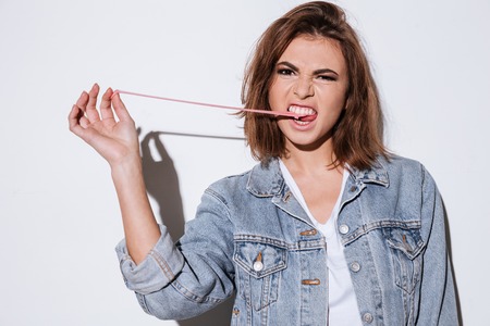 Image of a young lady dressed in jeans jacket standing isolated over white background stretching bubble gum. Look at camera.の写真素材