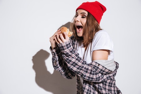 Image of hungry lady dressed in shirt in a cage print wearing hat standing isolated over white background and holding burgerの写真素材