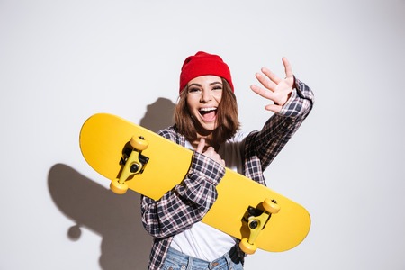 Picture of cheerful woman dressed in shirt in a cage print wearing hat standing isolated over white background holding skateboardの写真素材