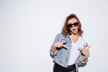 Photo of a young woman wearing sunglasses dressed in jeans jacket standing isolated over white background make peace gesture with mouth opened.の写真素材