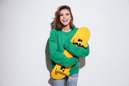 Photo of smiling skater woman dressed in green sweater standing isolated over white background with skateboard.の写真素材