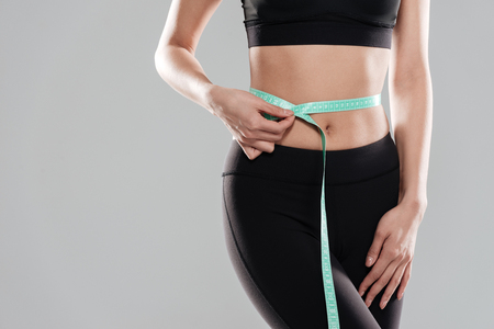 Closeup of woman athlete standing and using measuring tape on her waist over gray backgroundの写真素材