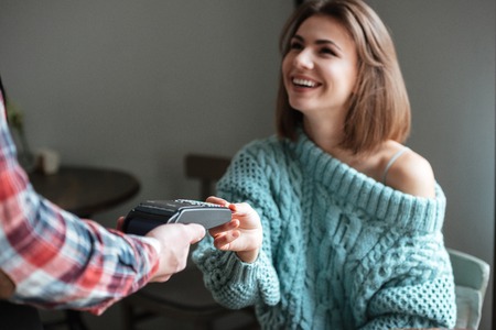 Image of beautiful young lady dressed in sweater sitting in cafe at cold winter day and pays for her order with debit card. Look aside.の写真素材