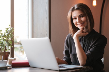 Picture of pretty young woman sitting at the table in cafe and using laptop computer. Look at camera.の写真素材