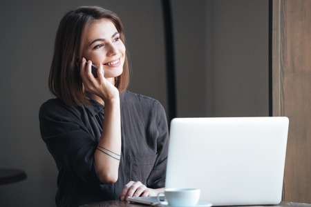 Photo of attractive woman sitting at the table in cafe and using laptop while talking by phone. Look aside.の写真素材